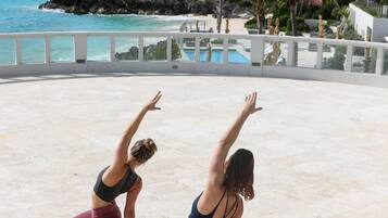 Dos personas practicando yoga en una playa, con vista al océano y un edificio en el fondo.