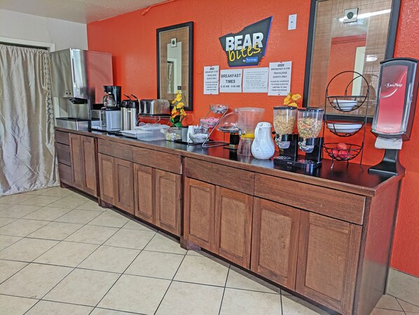 A breakfast bar with a sign reading "BEAR Bites by Tracloche," a coffee machine, a fruit bowl, and a juice dispenser. - Motel 6 Houston, TX – Hobby Airport (Houston)