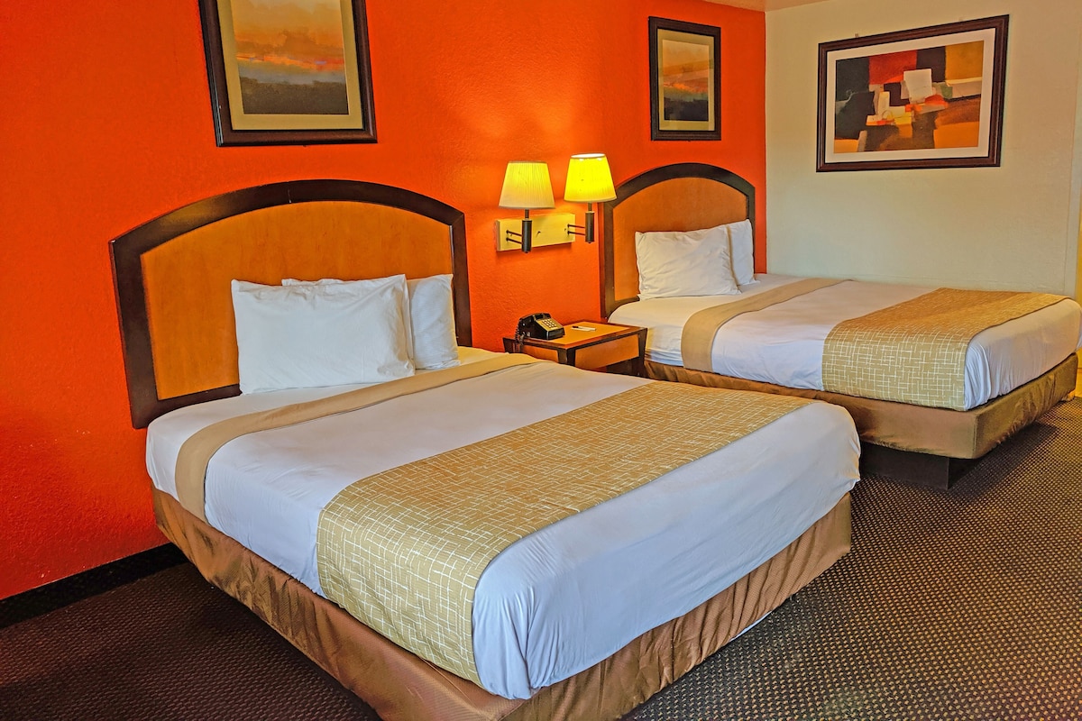 A hotel room with two beds, each with a wooden headboard, white bedding, and a beige throw blanket. There are two wall-mounted lamps on either side of the beds, and a painting above the left bed.