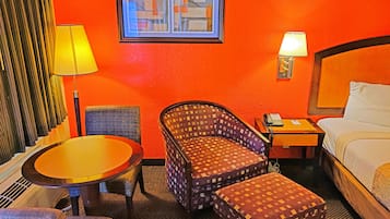 Una habitación de hotel con una cama, una mesa redonda, dos sillones, un pequeño reposapiés, una mesita de noche con un teléfono y un cuadro en la pared.