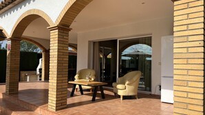 Een overdekt terras met een tafel en twee stoelen, een schuifdeur en een koelkast.