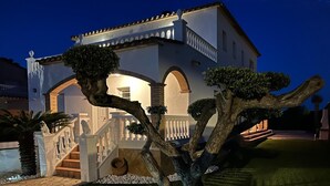 Een goed verlicht huis met twee verdiepingen, een witte gevel, een balkon en een stenen trap die naar de ingang leidt.