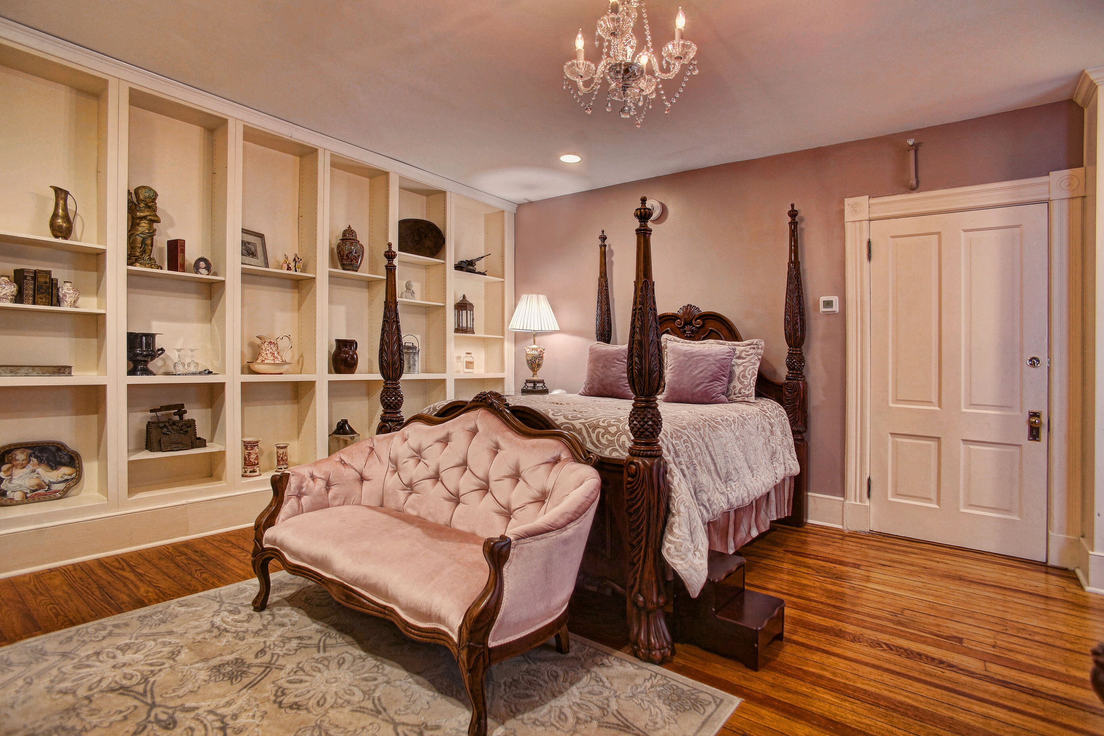 A bedroom with a four-poster bed, a pink armchair, and built-in shelving with various decorative items.