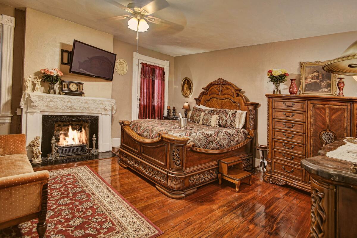 A traditional bedroom with a fireplace, a large bed, a wooden dresser, and a television mounted on the wall.