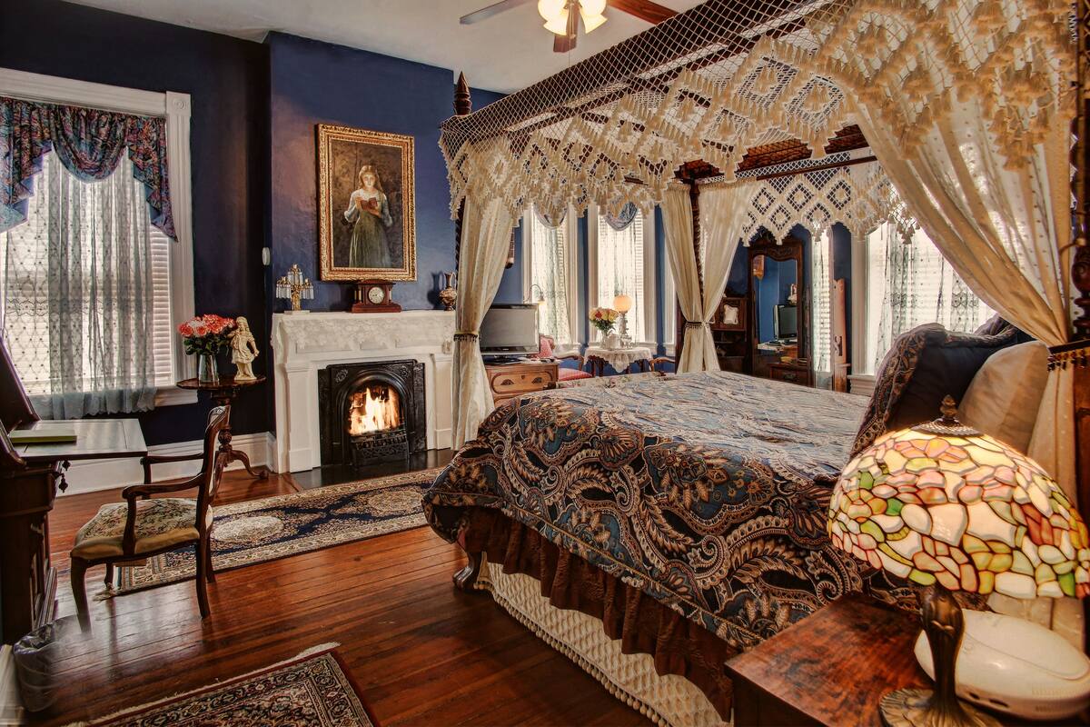 A bedroom with a large bed, a fireplace, a chandelier, and a view of the outdoors.