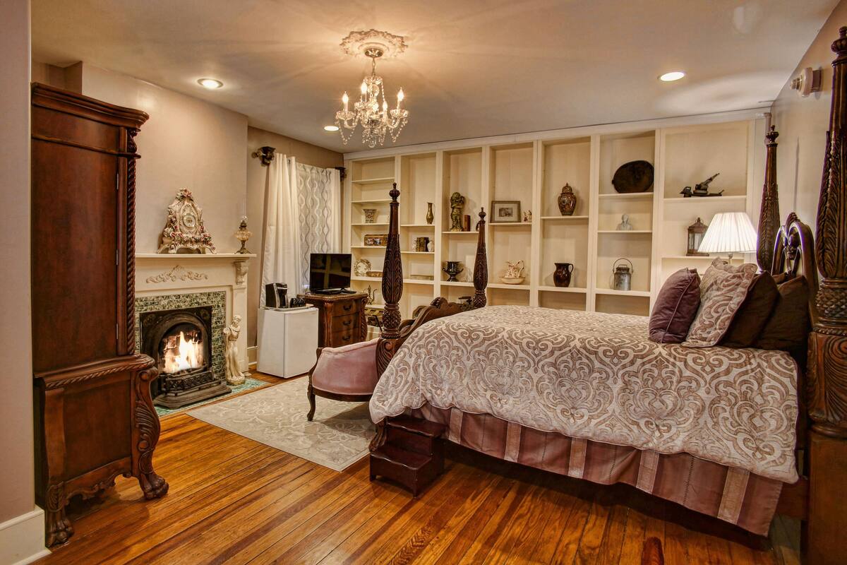 A bedroom with a large bed, a fireplace, a chandelier, and a bookshelf.