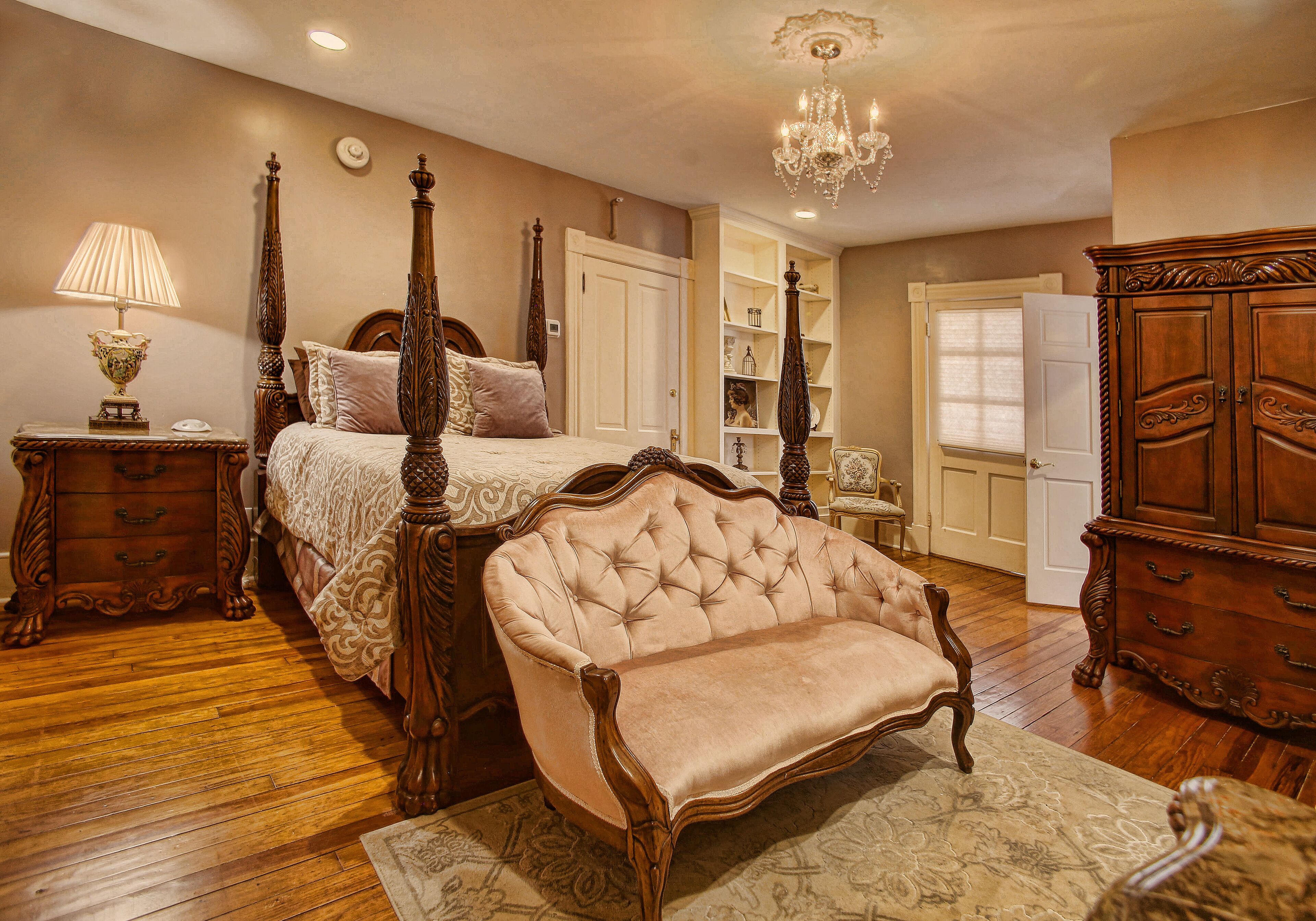 A four-poster bed with a tufted headboard, a chaise lounge, and a wooden dresser in a room with wooden flooring and a chandelier.