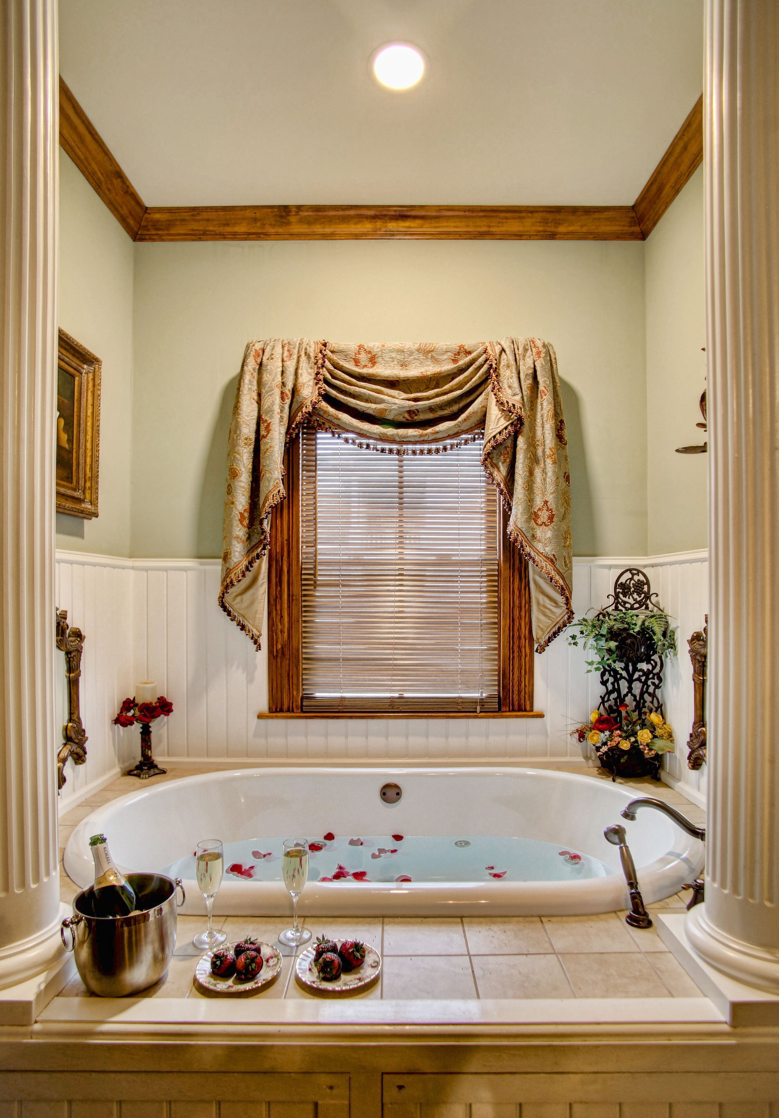A bathroom with a bathtub, a window with curtains, a vase with flowers, a bottle of champagne, and two plates with strawberries.