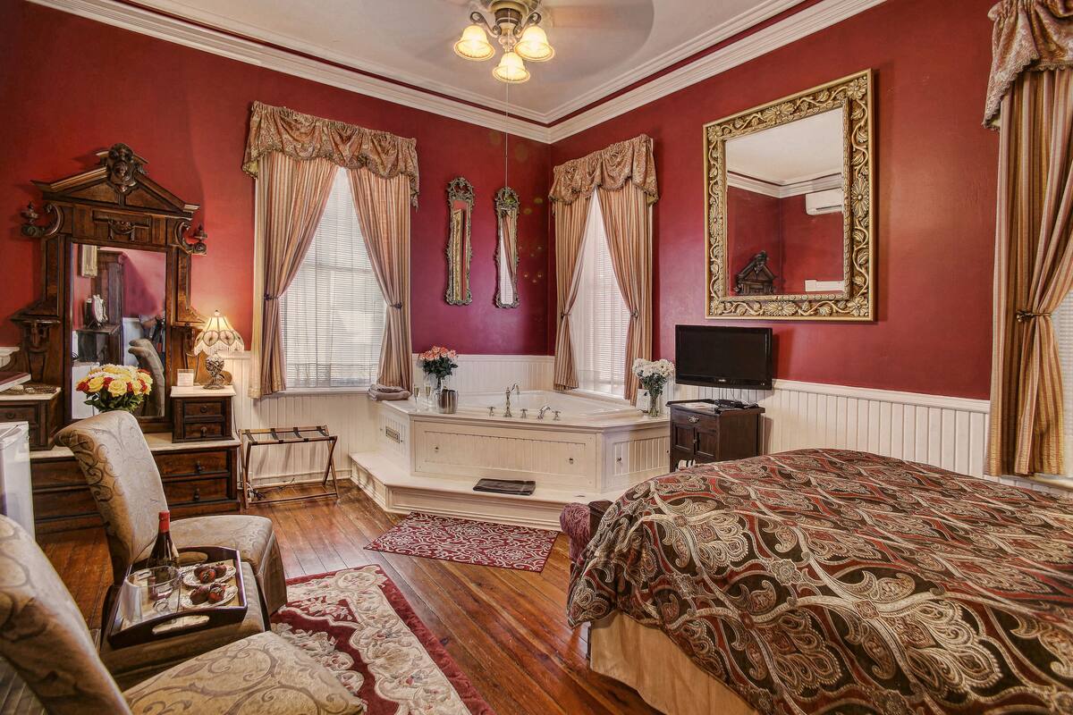 A bedroom with a large bed, a sitting area with a chair, a vanity with a mirror, and a bathtub.