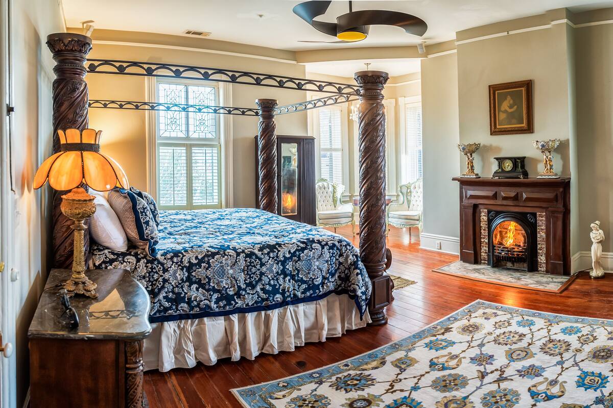 A bedroom with a four-poster bed, a fireplace, and a large window with shutters.