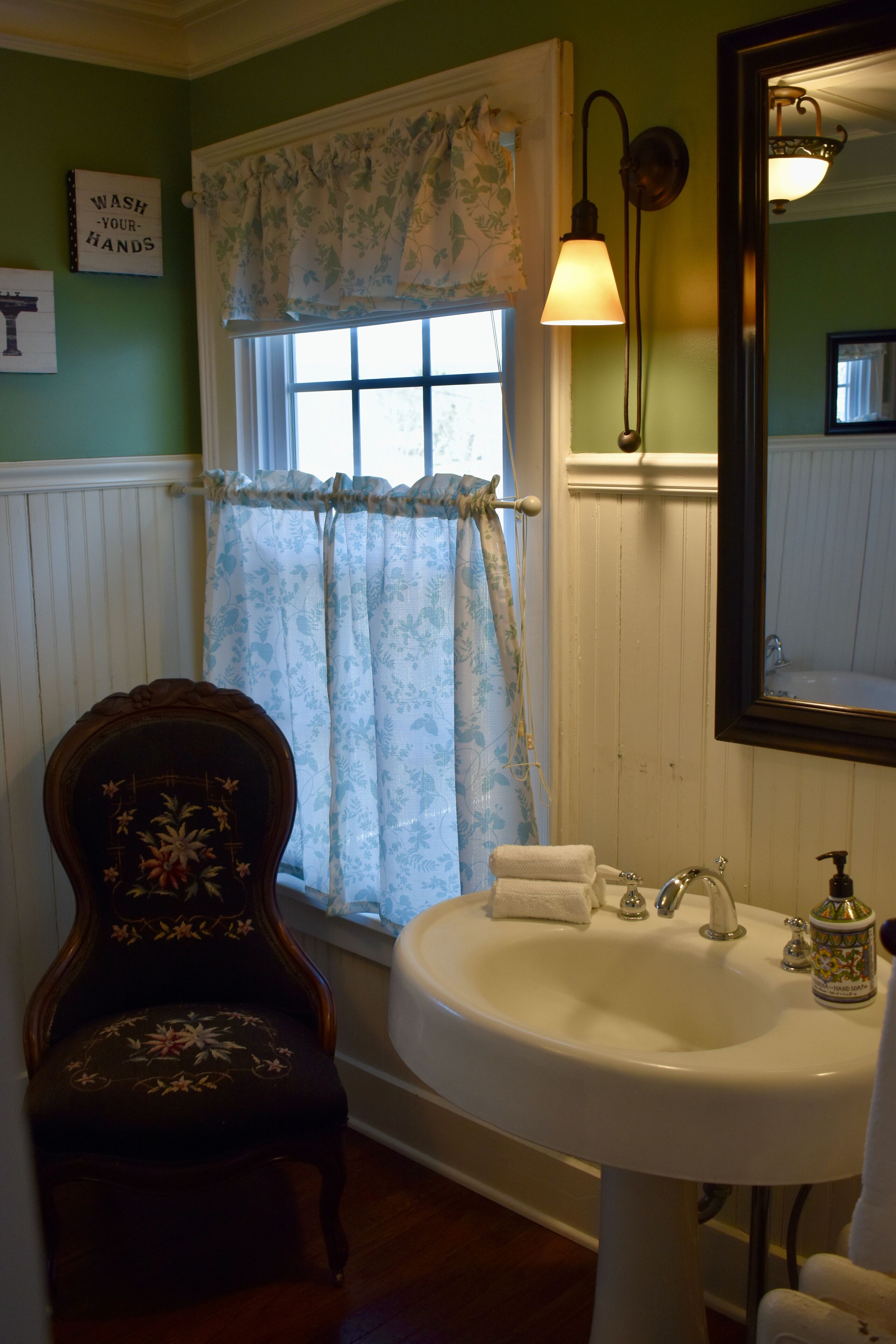 A bathroom with a white sink, a mirror, a floral-patterned chair, and a window with a curtain.