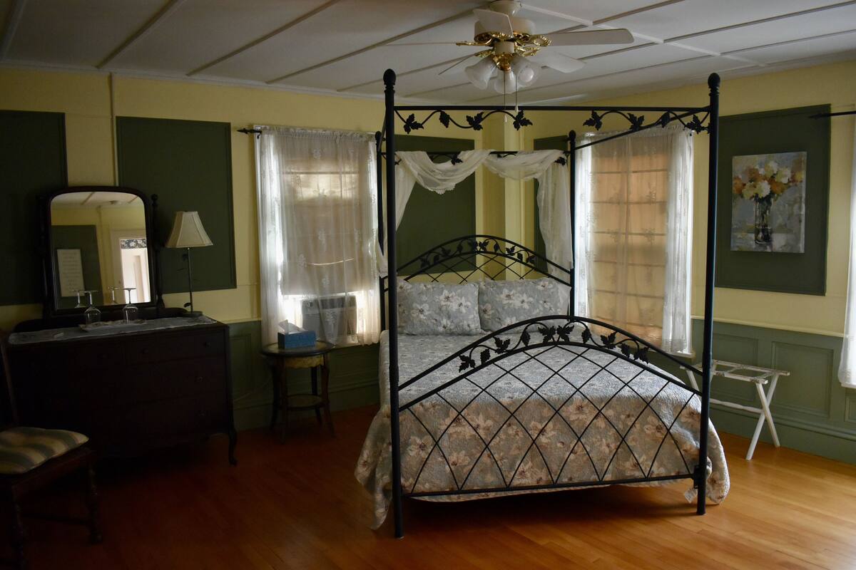 A four-poster bed with a floral bedspread, a dresser with a mirror, and a small table with a lamp.
