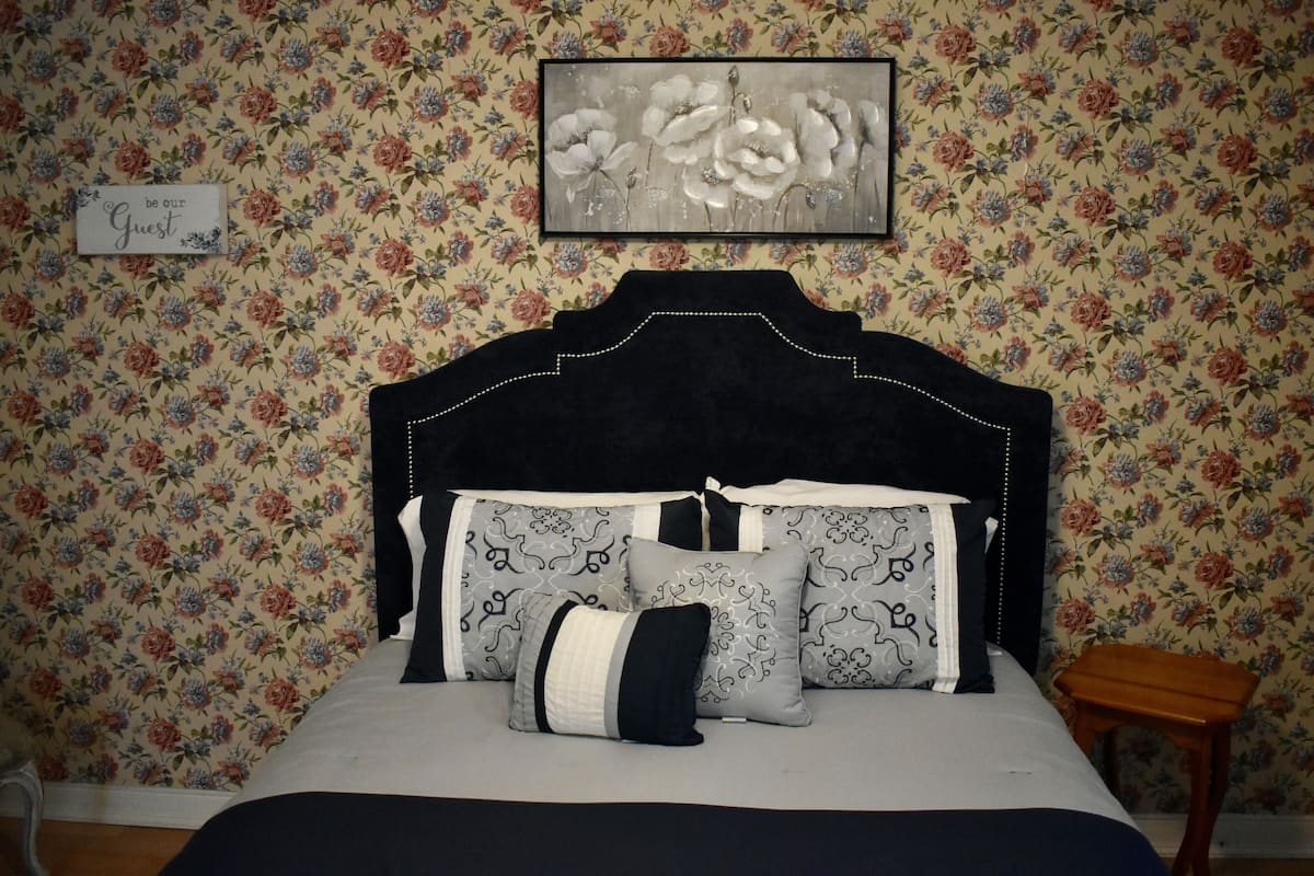 A bedroom with floral wallpaper, a black upholstered headboard, and a framed floral painting above the bed.