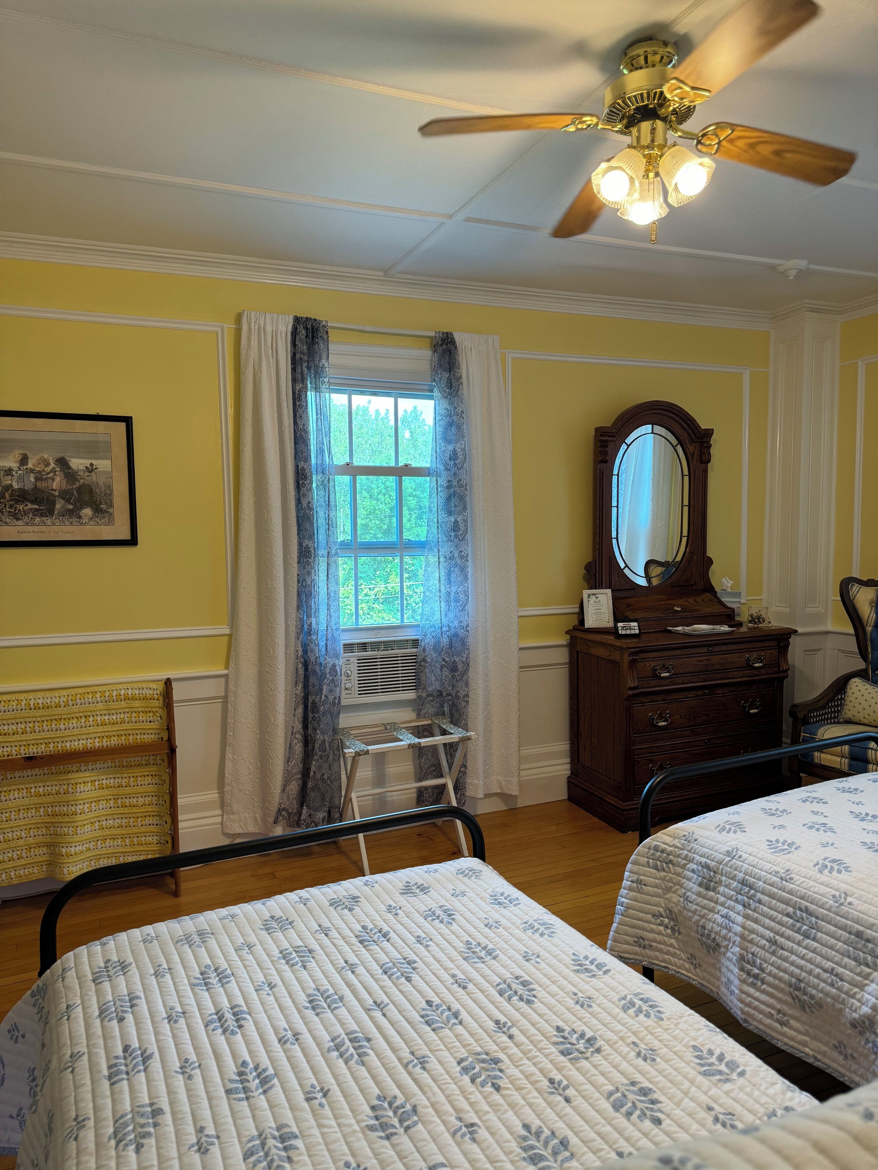 Une chambre avec deux lits, un ventilateur de plafond, une fenêtre avec des rideaux, une commode et un tableau encadré au mur.
