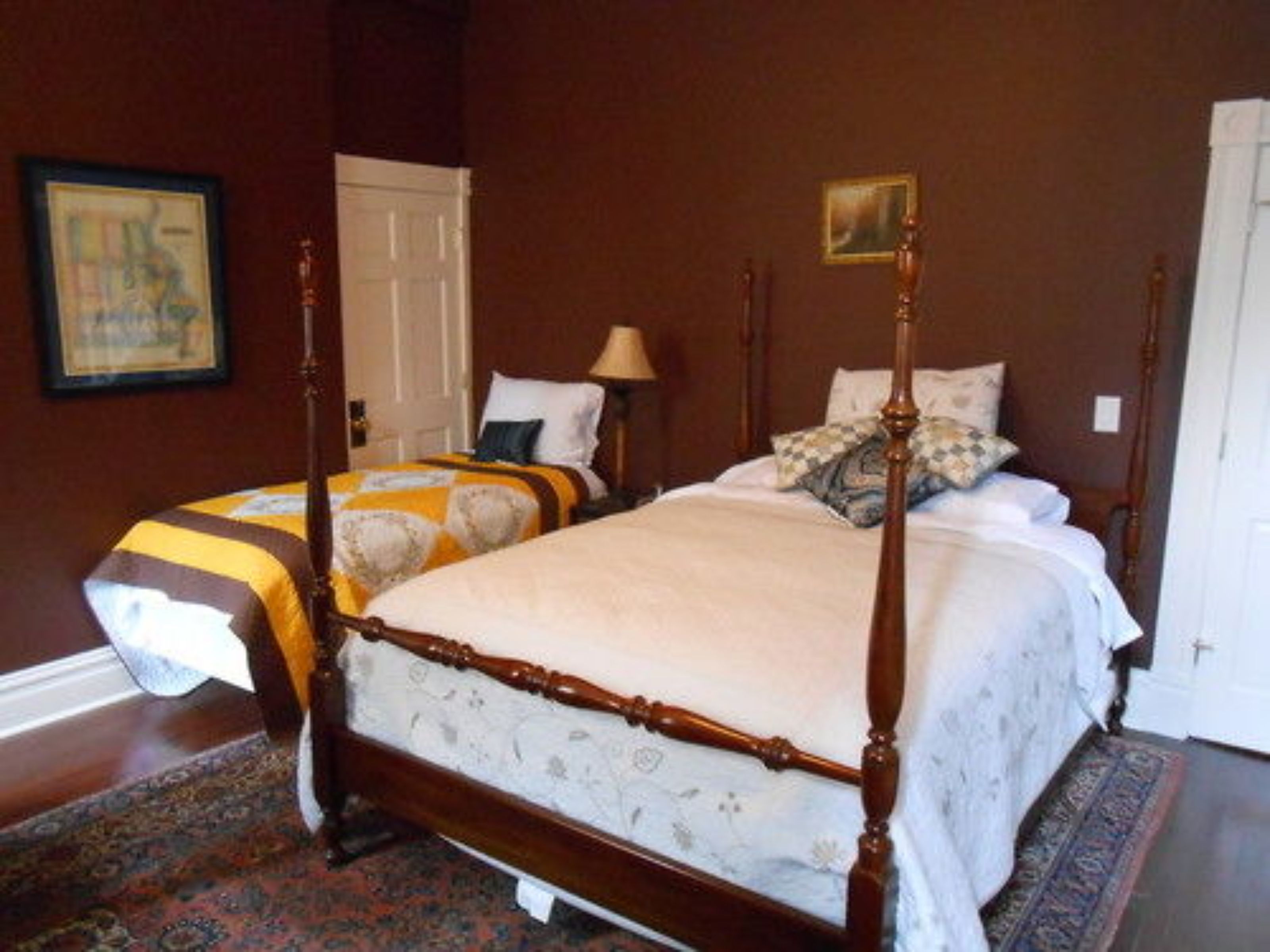 A traditional four-poster bed with a wooden frame and a floral bedspread. There is a nightstand with a lamp and a framed picture on the wall.