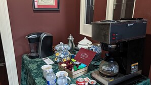 Una estación de café con una cafetera Bunn, una jarra de café, un soporte para tazas y varias cápsulas de café. También hay agua embotellada, una lata de Coca-Cola, una lata de cerveza Budweiser y una caja roja con la etiqueta 'Bennett's'.