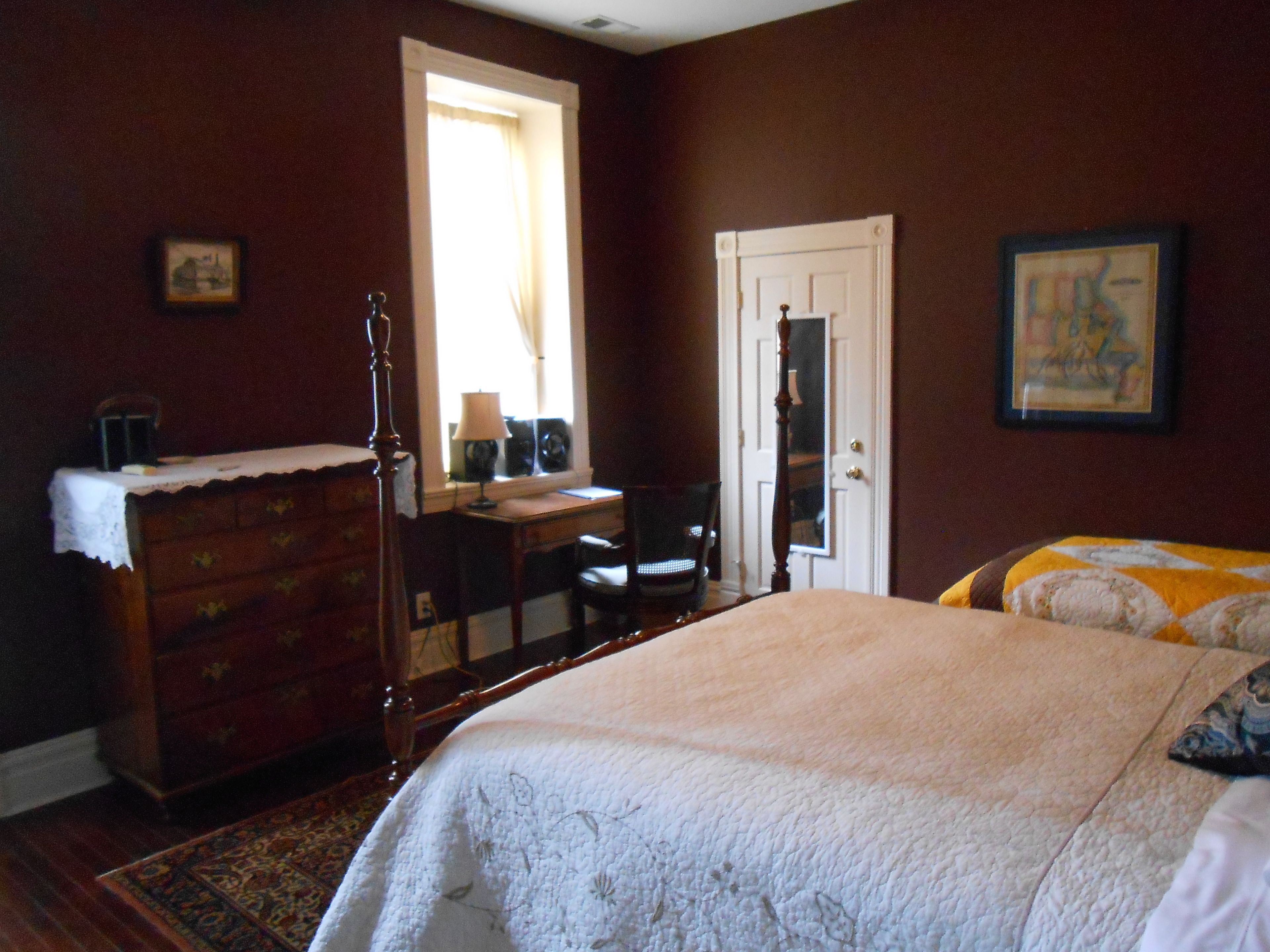 Quarto com cama grande, cômoda, escrivaninha e uma cadeira. Há uma janela com cortinas, uma porta e um quadro na parede.