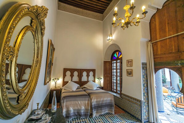 A bedroom with a large ornate mirror, a bed with two pillows, a nightstand with a bottle, and a chandelier.
