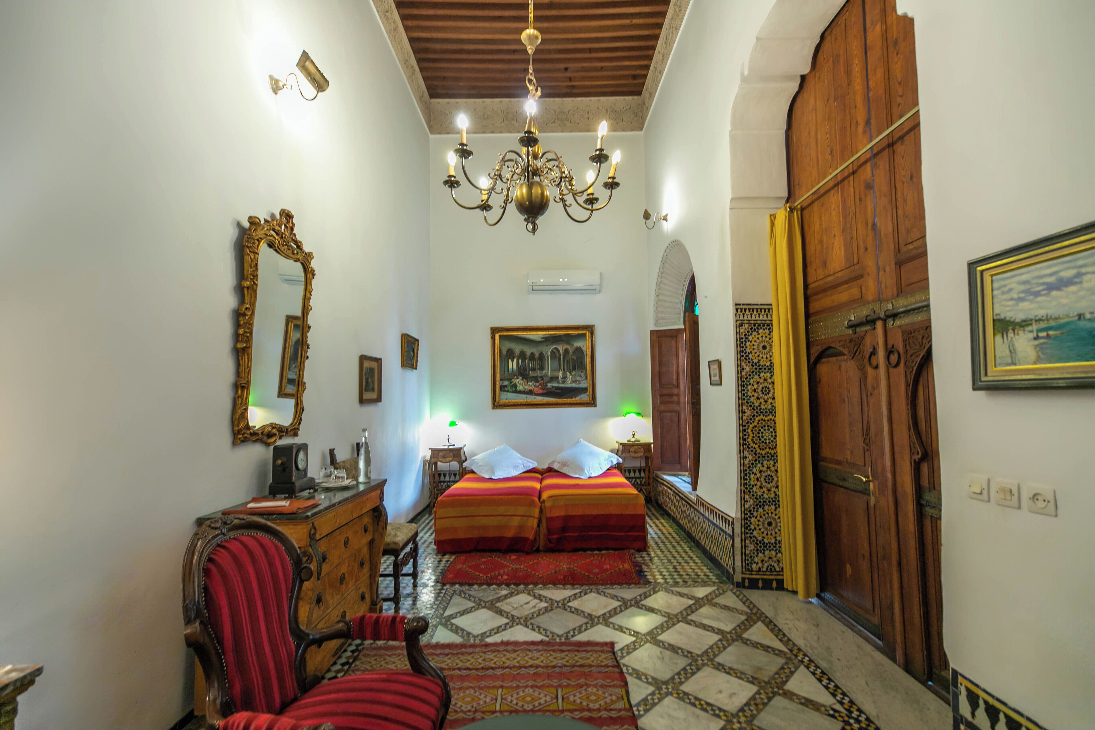 A room with a chandelier, a large mirror, two beds with red and orange bedding, a wooden dresser, and a patterned floor.