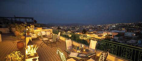 A rooftop terrace with a table and chairs, potted plants, and a view of a cityscape at night.