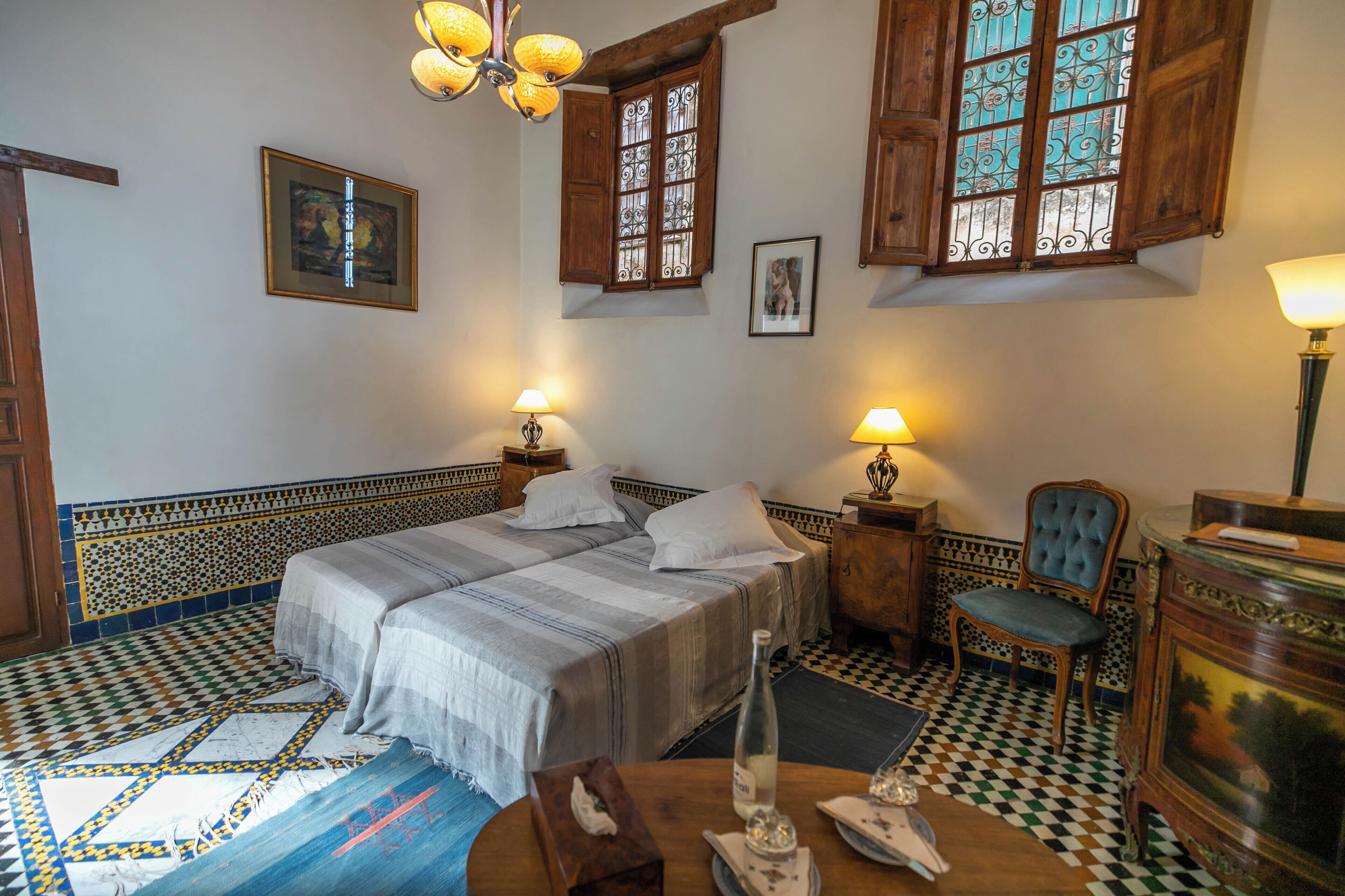 Una habitació amb un llit, dues taules de nit, una cadira, una taula amb una ampolla i gots, i una làmpada.