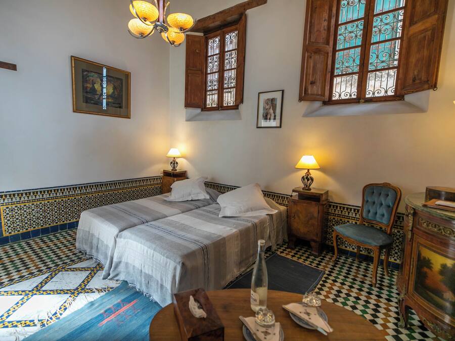 Una habitació amb un llit, dues taules de nit, una cadira, una taula amb una ampolla i gots, i una làmpada.