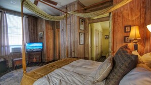 A room with a large bed, a television, wooden walls, and a window with curtains. - GardenSong B&B (Natchez)