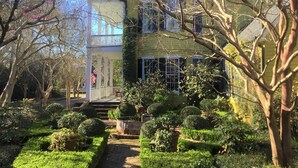 A two-story house with a covered porch, a flag, and a well-maintained garden with various shrubs and a brick pathway. - GardenSong B&B (Natchez)