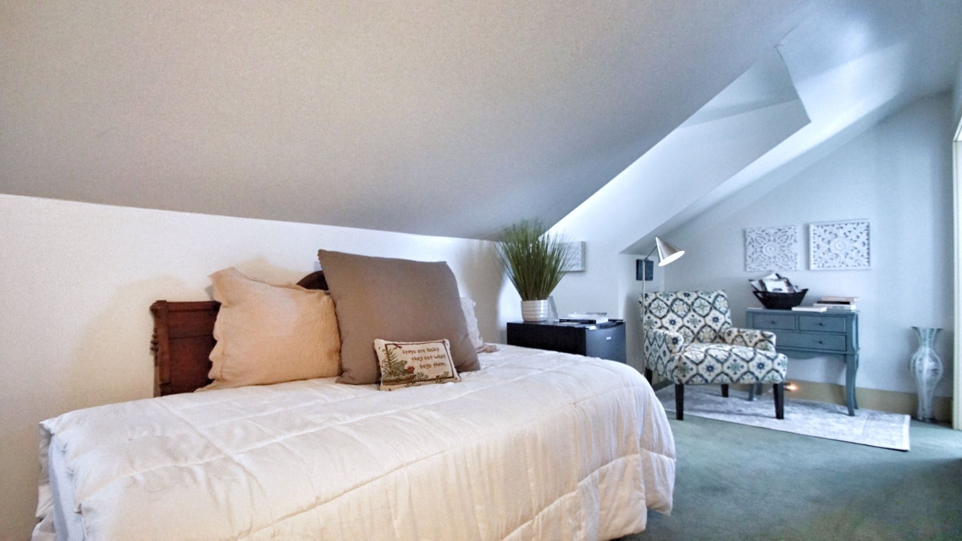 Une chambre à coucher avec un lit, des oreillers, un bureau, une chaise, une plante et des œuvres d’art accrochées aux murs.