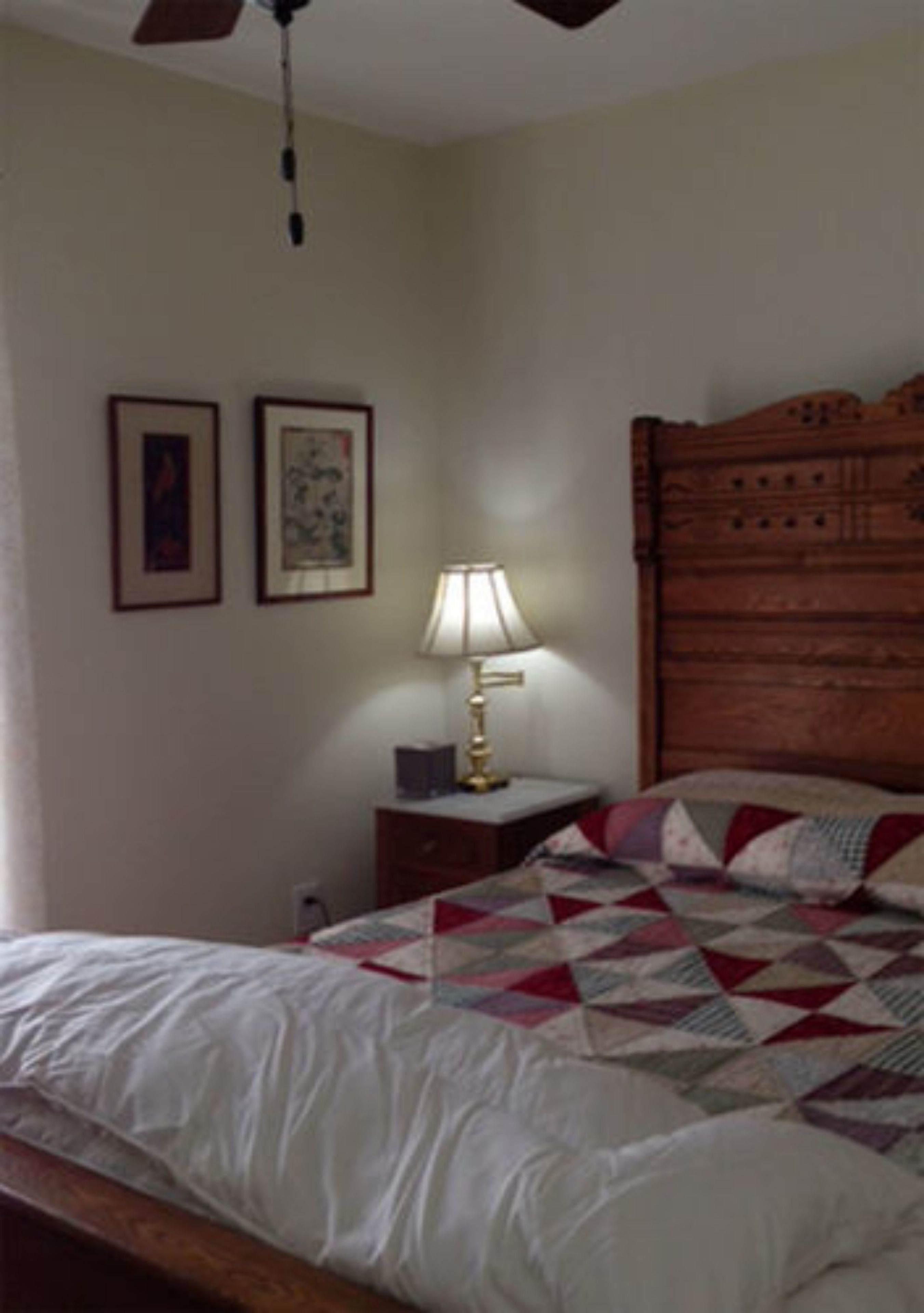 A bedroom with a wooden headboard, a bed with a quilt, a nightstand with a lamp, and two framed pictures on the wall.