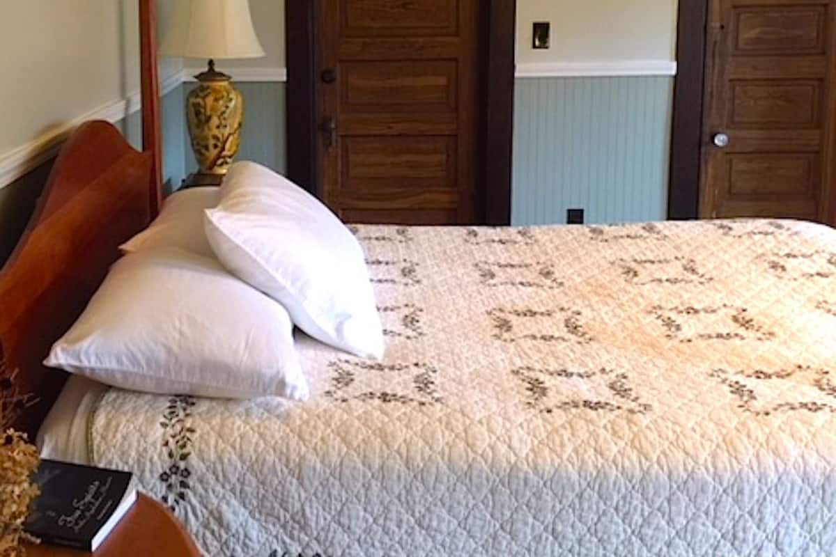A bedroom with a wooden bed frame, a quilted bedspread, two white pillows, a bedside table with a lamp, and two wooden doors.