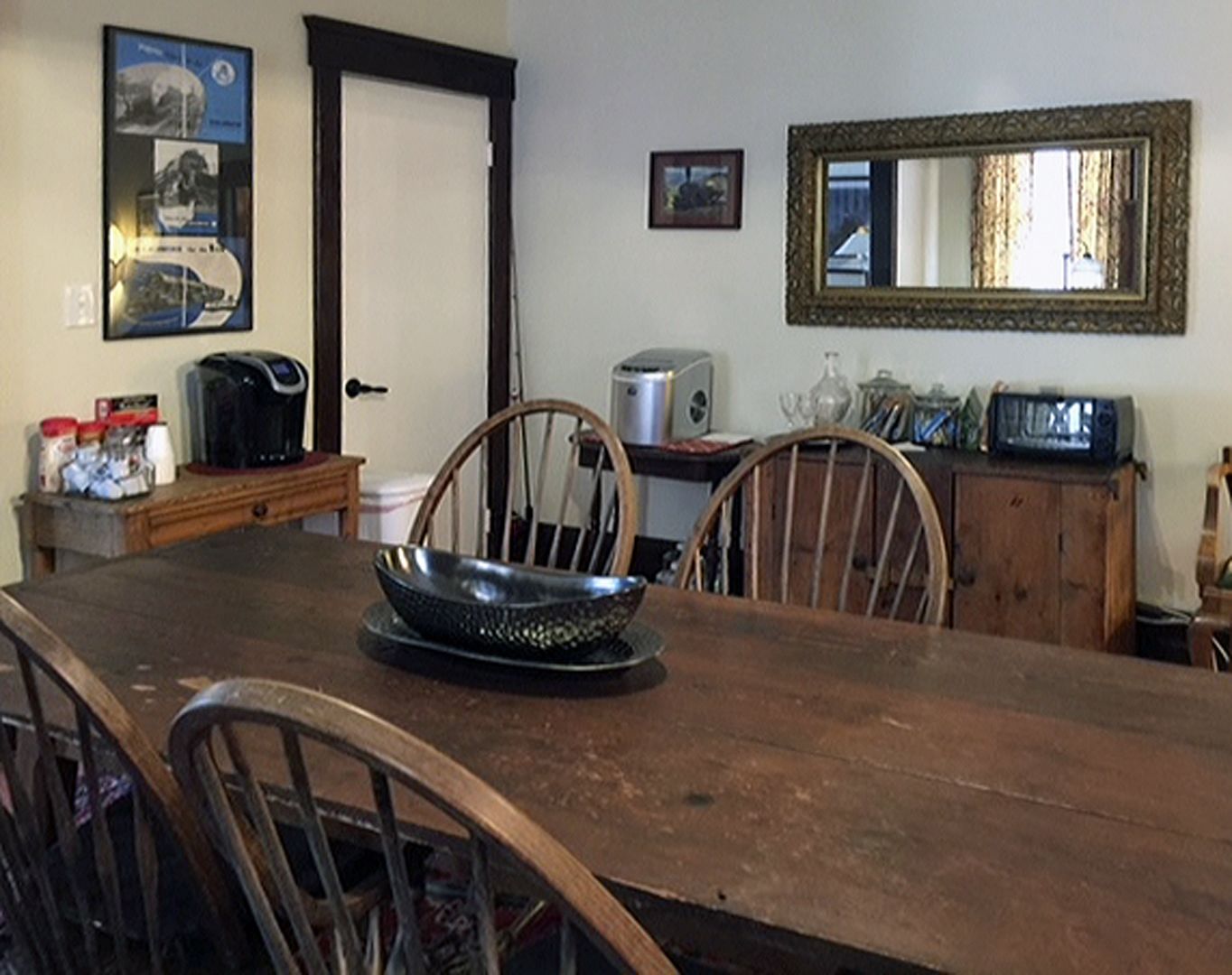 A dining area with a wooden table, chairs, a coffee maker, a radio, and a mirror on the wall.