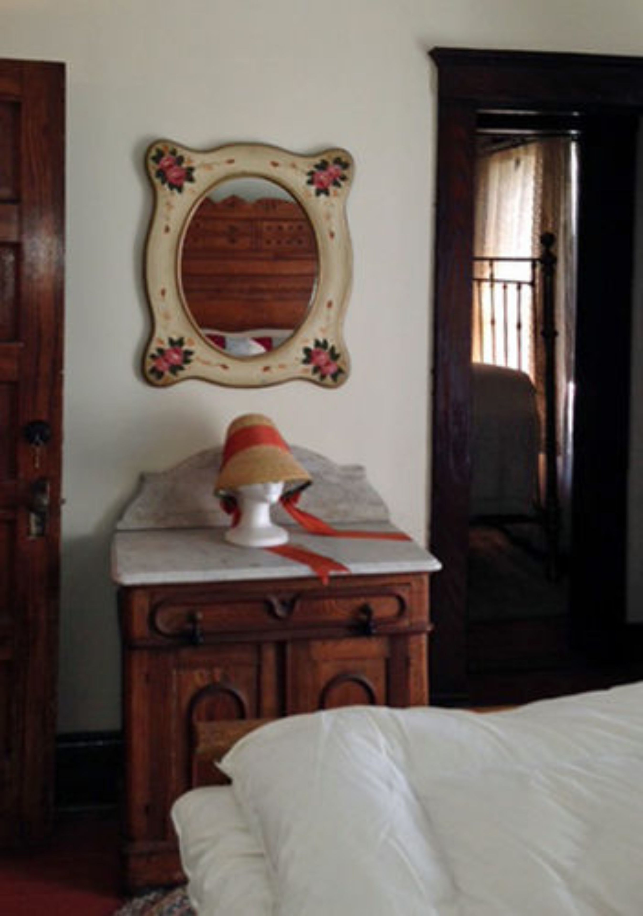 Un dormitorio con un aparador de madera, un espejo con diseño floral y una cama con ropa blanca.