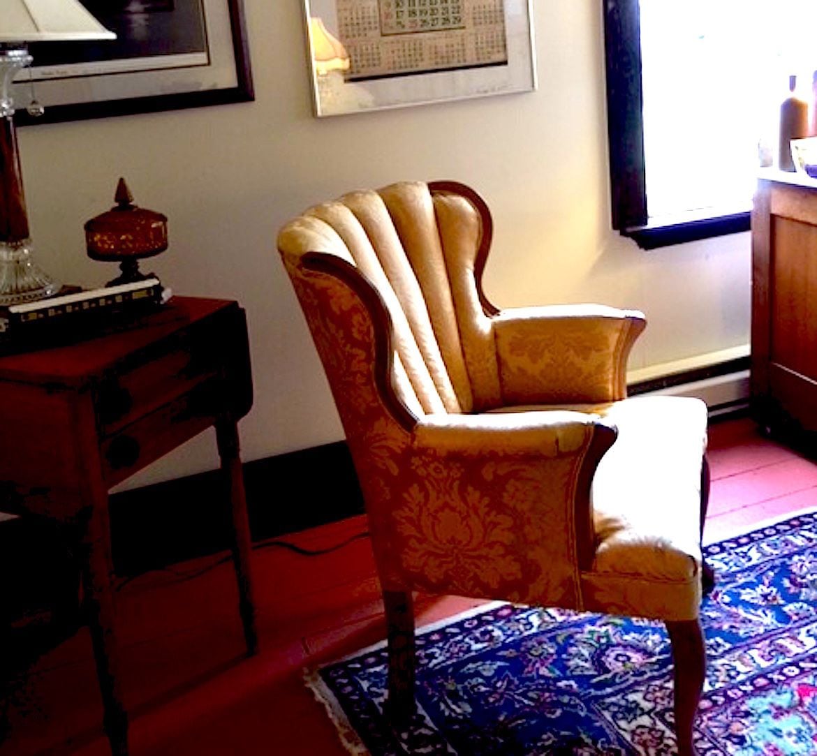 Une pièce comprenant un fauteuil à motifs, une petite table en bois avec une lampe et un tableau encadré accroché au mur.