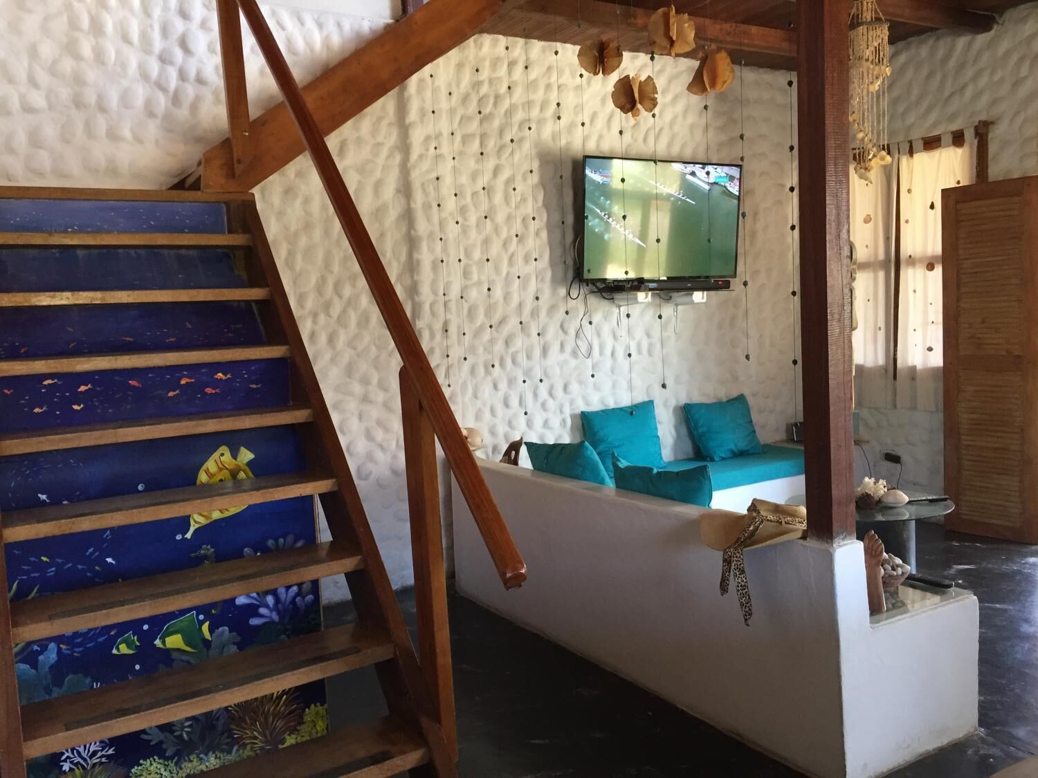 Una escalera con peldaños azules y un mural de peces, una sala de estar con un sofá color turquesa y un televisor en la pared, y una estructura de techo de madera.