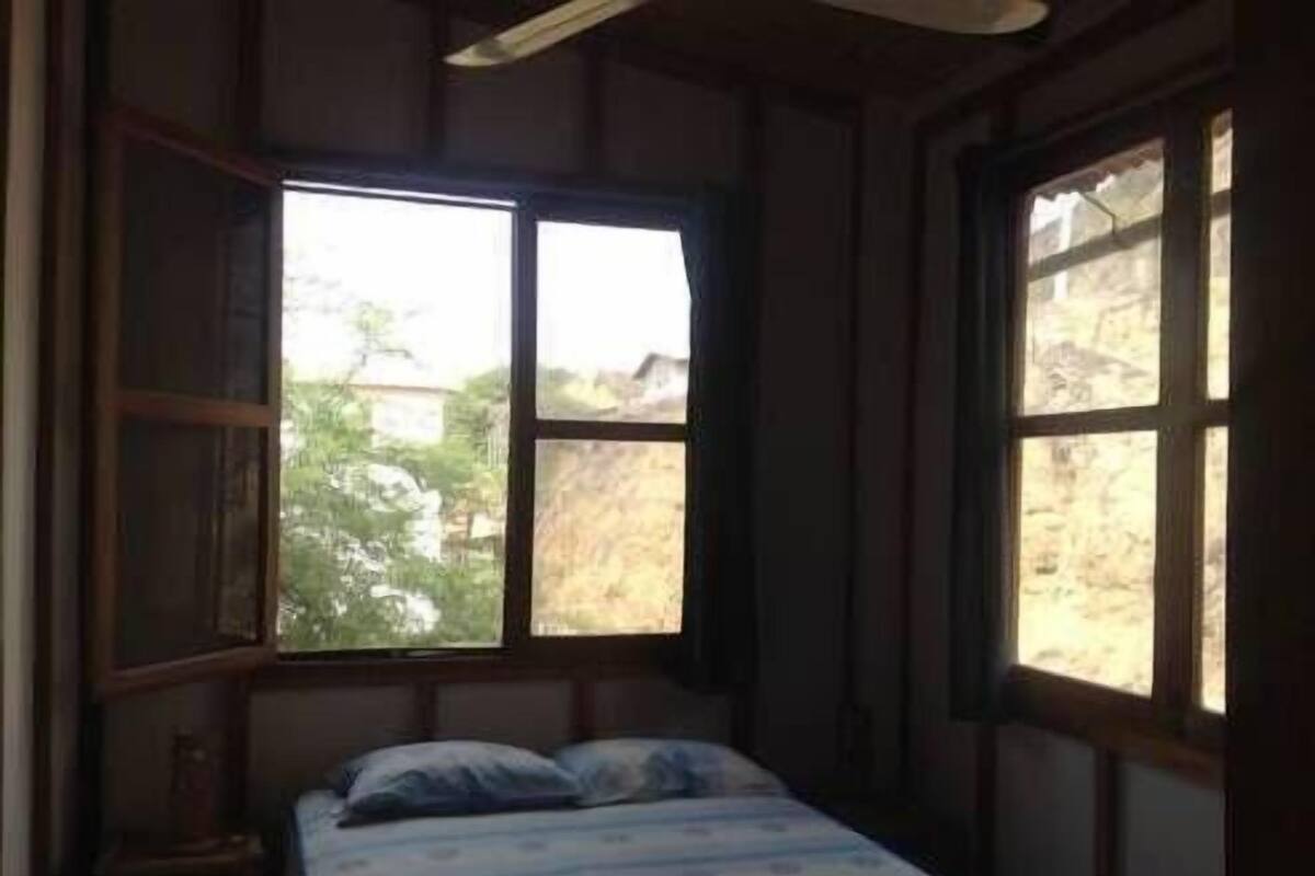 Une petite pièce peu éclairée, avec un lit simple, de grandes fenêtres et un ventilateur au plafond.