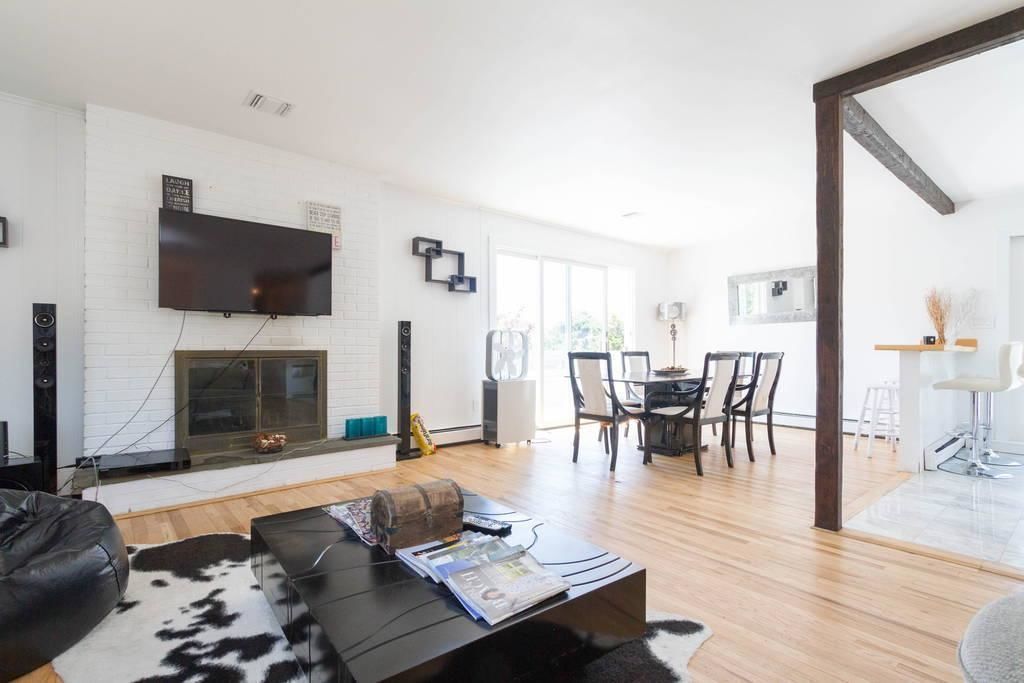 A modern living room with a fireplace, a flat-screen TV mounted on the wall, a dining area with a table and chairs, and a kitchen area visible through an open door.