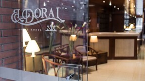 Zona de recepción de un hotel con un ventanal que muestra el nombre 'DORAI APARTMENT HOTEL'. En el interior, hay sillas de madera, una mesa y una lámpara.
