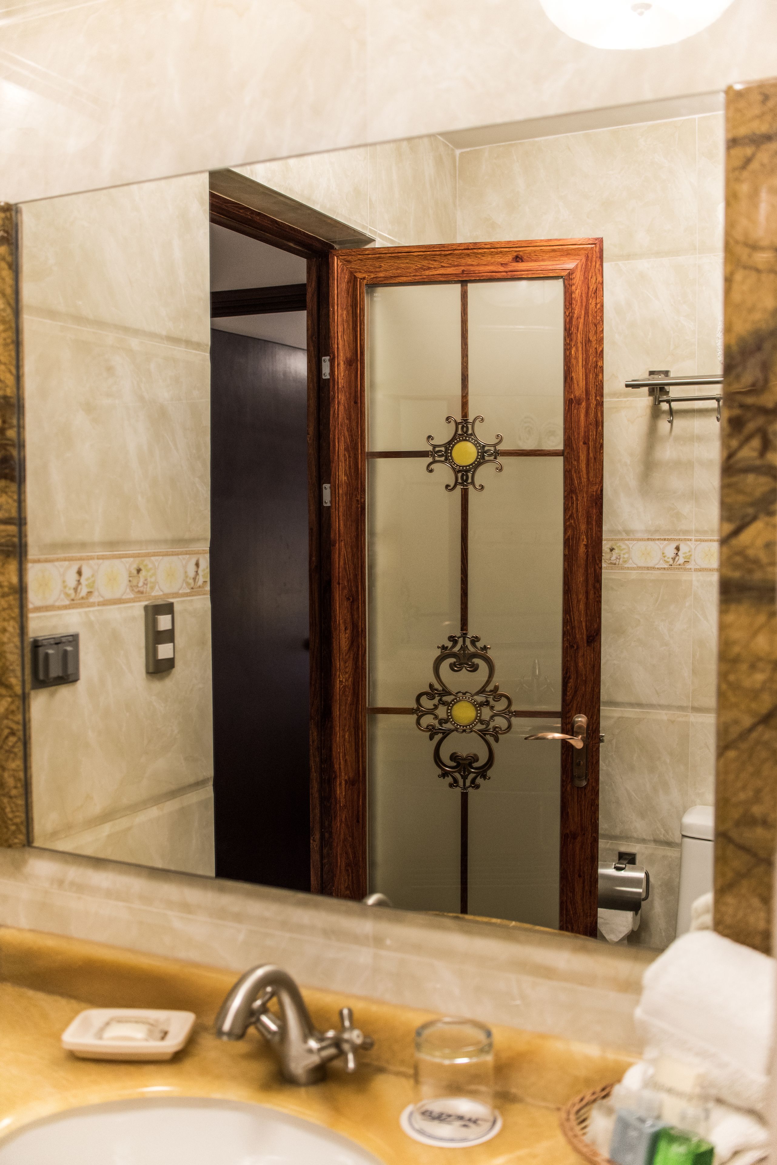 A bathroom with a marble wall, a wooden door with a glass panel, a sink with a faucet, and a mirror reflecting the door.