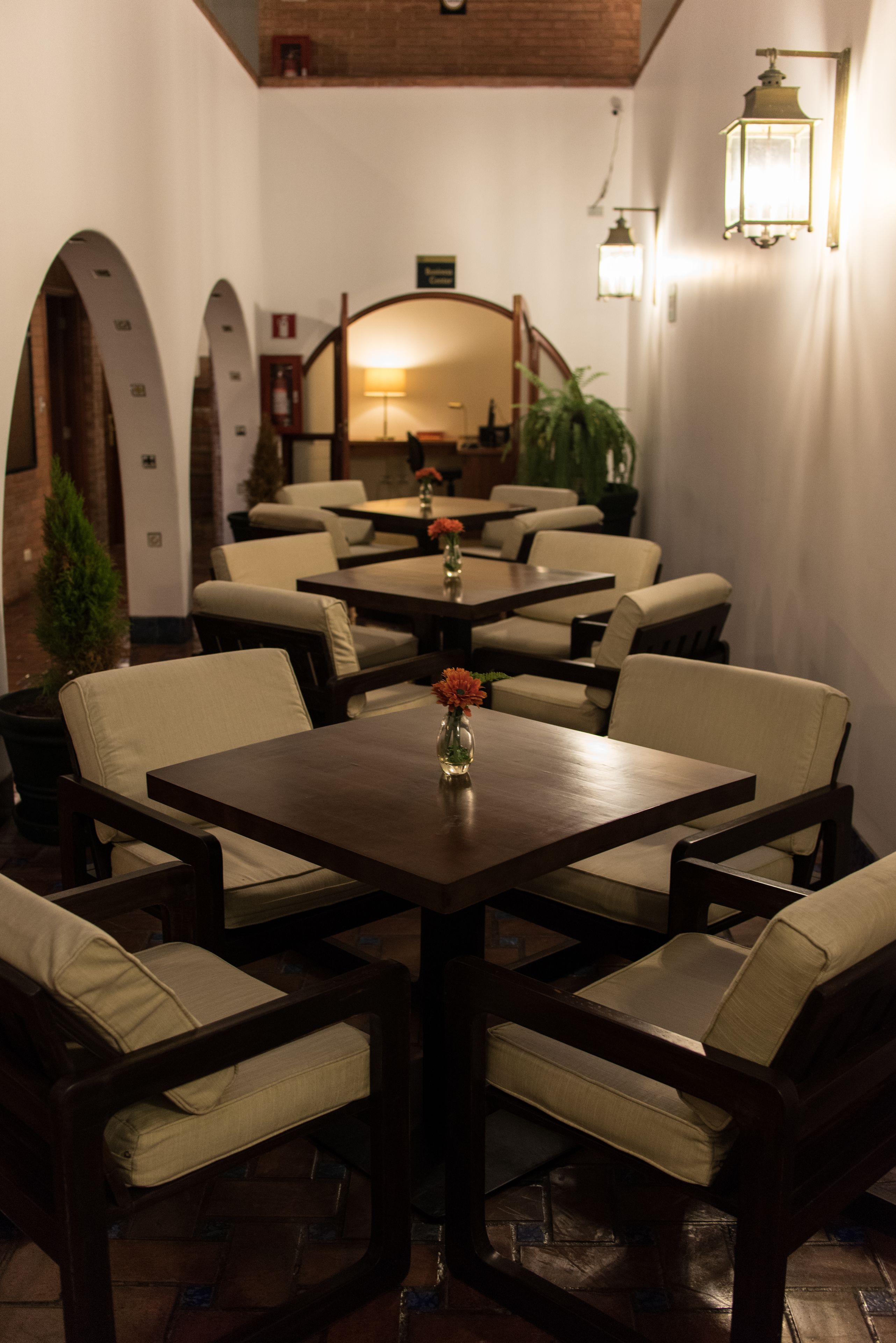A dining area with a central table surrounded by cushioned chairs, a vase with flowers, and a brick wall with an arched doorway.