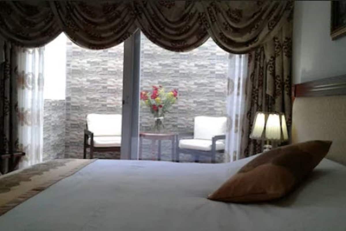 Habitación de hotel con cama, ventana con cortinas, una mesita con flores y dos sillas.