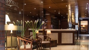 Vestíbulo de hotel con mostrador de recepción, sillas de madera, un jarrón con flores blancas y una pared de ladrillo.