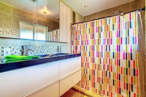 Un baño moderno con zona de ducha azulejada, un espejo grande y un mostrador negro con una toalla verde.