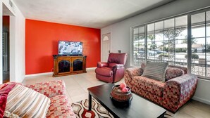 A living room with a red accent wall, a television on a wooden cabinet, a red leather chair, a patterned sofa, and a coffee table with decorative items.