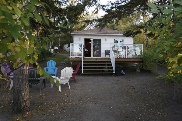 A small white cabin with a porch, surrounded by trees. There are several chairs and a table on the porch. - Lakefront Cottage in Manitou Beach (Manitou Beach)