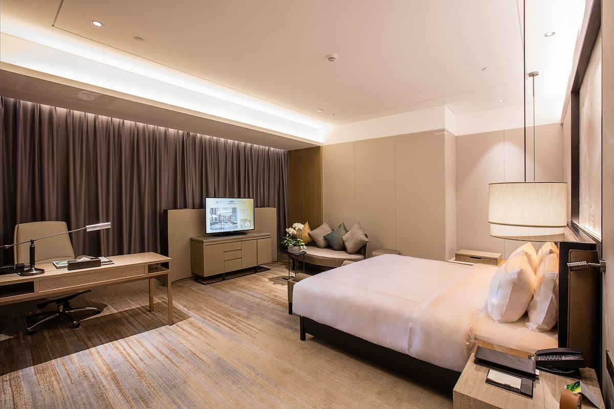 Quarto de hotel moderno com uma cama grande, uma escrivaninha com computador, uma televisão e uma mesa de cabeceira com abajur.