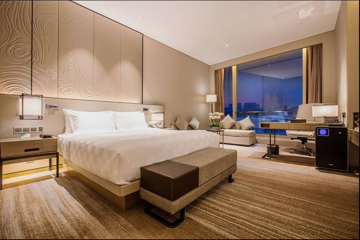 Quarto de hotel moderno com uma cama grande, uma escrivaninha e vista para a paisagem urbana.
