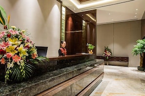 A reception area with a marble counter, a woman standing behind it, and a large floral arrangement on the left. - Golden Eagle Summit Hotel Nanjing China (Nanjing)