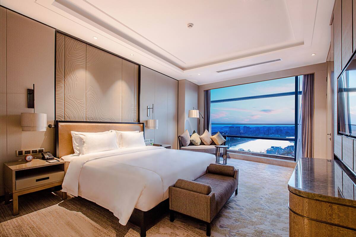 Quarto de hotel moderno com uma cama grande, mesa de cabeceira, escrivaninha e vista para a cidade através de amplas janelas.