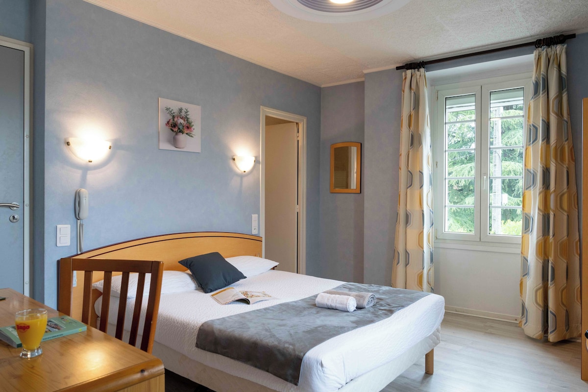 Ett hotellrum med en säng, en trästol, ett bord med ett glas apelsinjuice och ett fönster med gardiner.